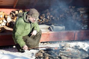A man cooks sausages over a wood fire. Behind him is a lean-to wilderness shelter filled with fire wood. These shelters are common in Finnish Lapland to allow travellers to warm themselves and make food and drinks in the freezing arctic winter.